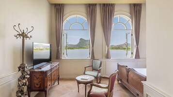 A room with a TV, a wooden cabinet, two chairs, a round table, and large windows with curtains.