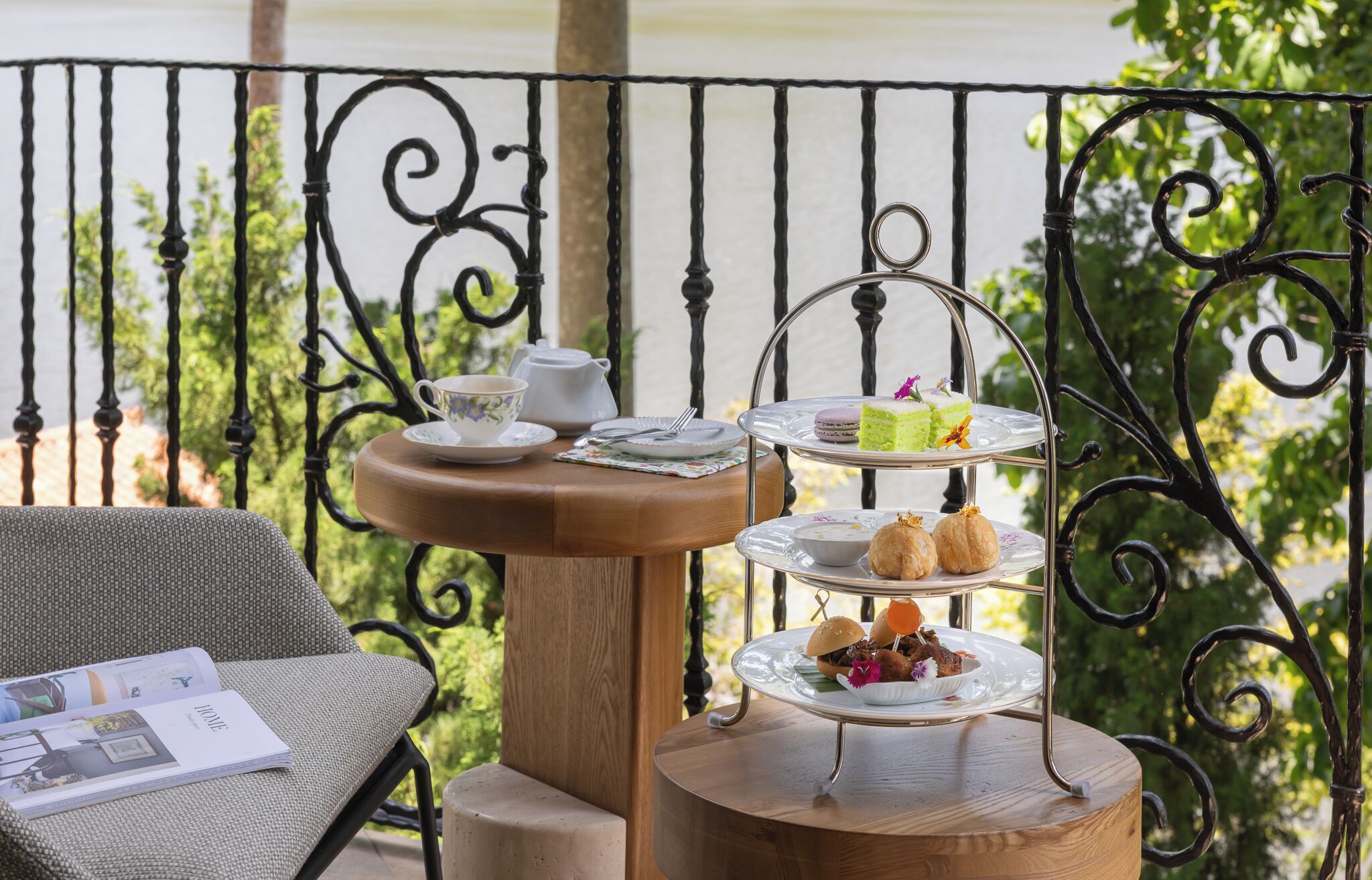 Un elegante soporte de tres niveles para servir té, con diversos pasteles y una tetera sobre una mesa de madera, ubicado en un balcón con barandilla de hierro forjado.