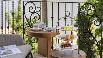 Un elegante soporte de tres niveles para servir té, con diversos pasteles y una tetera sobre una mesa de madera, ubicado en un balcón con barandilla de hierro forjado.