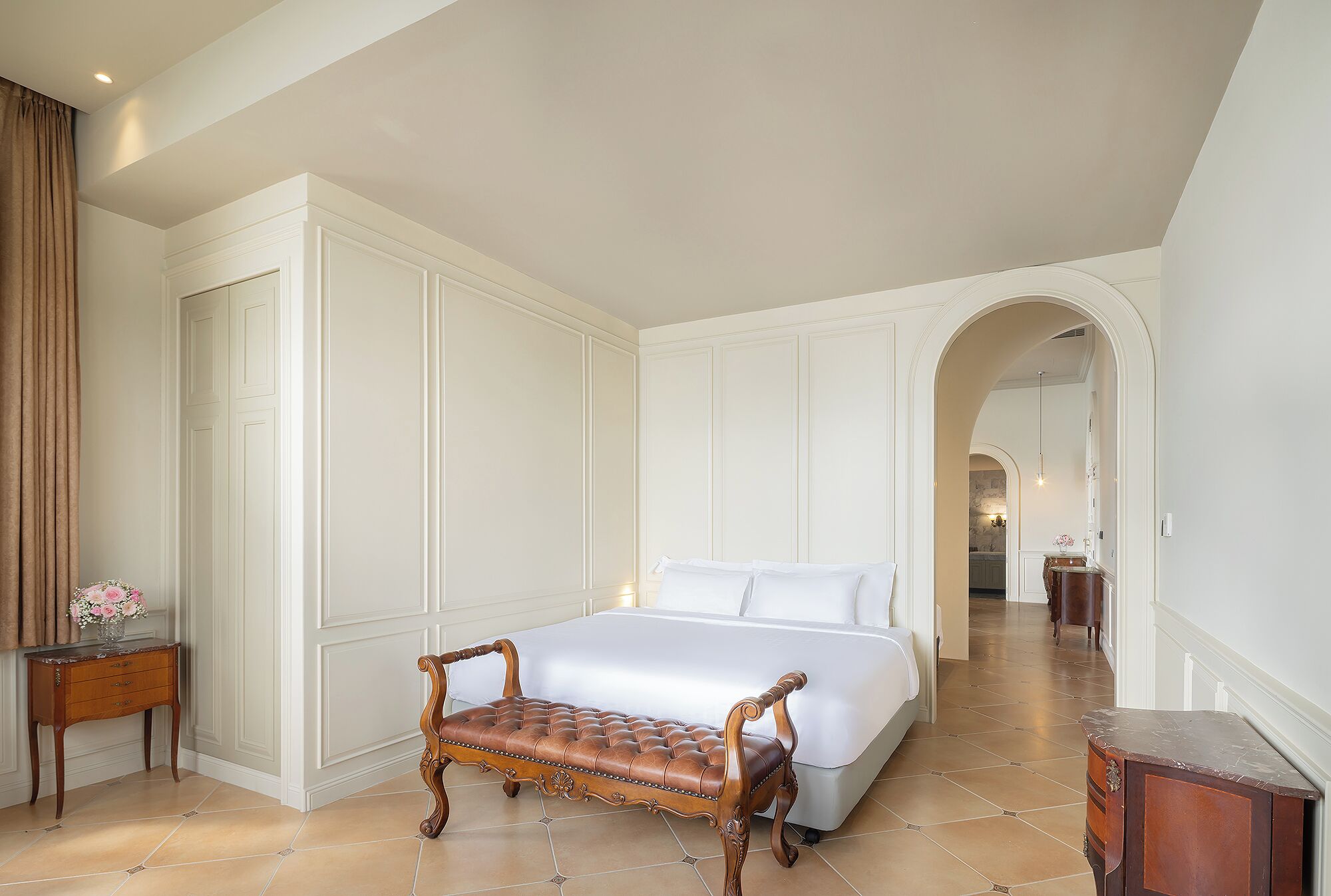 Un dormitorio de hotel con una cama grande, un banco de madera, una mesita de noche y un baño visible a través de un arco.