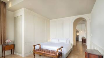 Un dormitorio de hotel con una cama grande, un banco de madera, una mesita de noche y un baño visible a través de un arco.