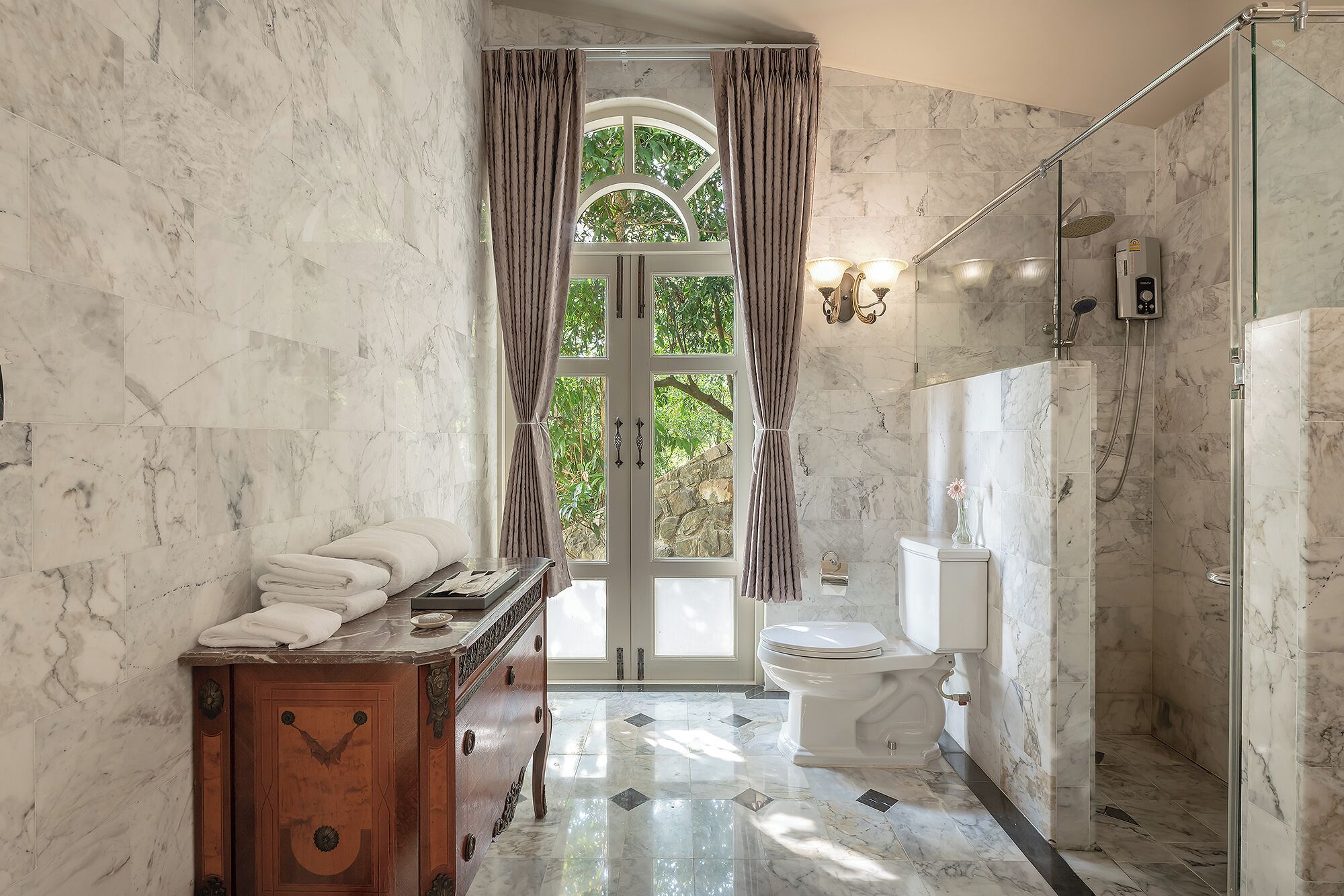Un baño con paredes de mármol, tocador de madera, inodoro y zona de ducha.