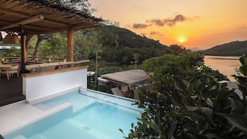 Ein Schwimmbecken mit Blick auf einen Sonnenuntergang über dem Wasser, umgeben von üppiger Vegetation und einer Holzkonstruktion.