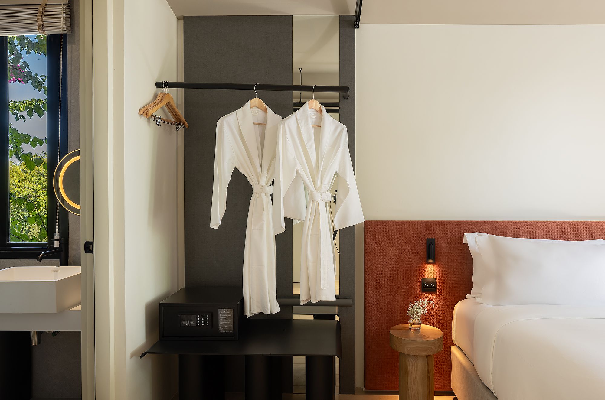 A hotel room with a bed, a bedside table with a lamp, a wall-mounted TV, and a clothing rack with two white robes.