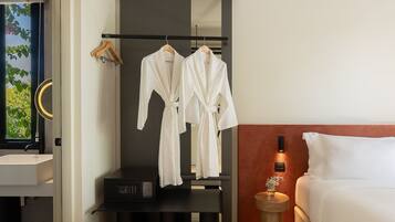 A hotel room with a bed, a bedside table with a lamp, a wall-mounted TV, and a clothing rack with two white robes.