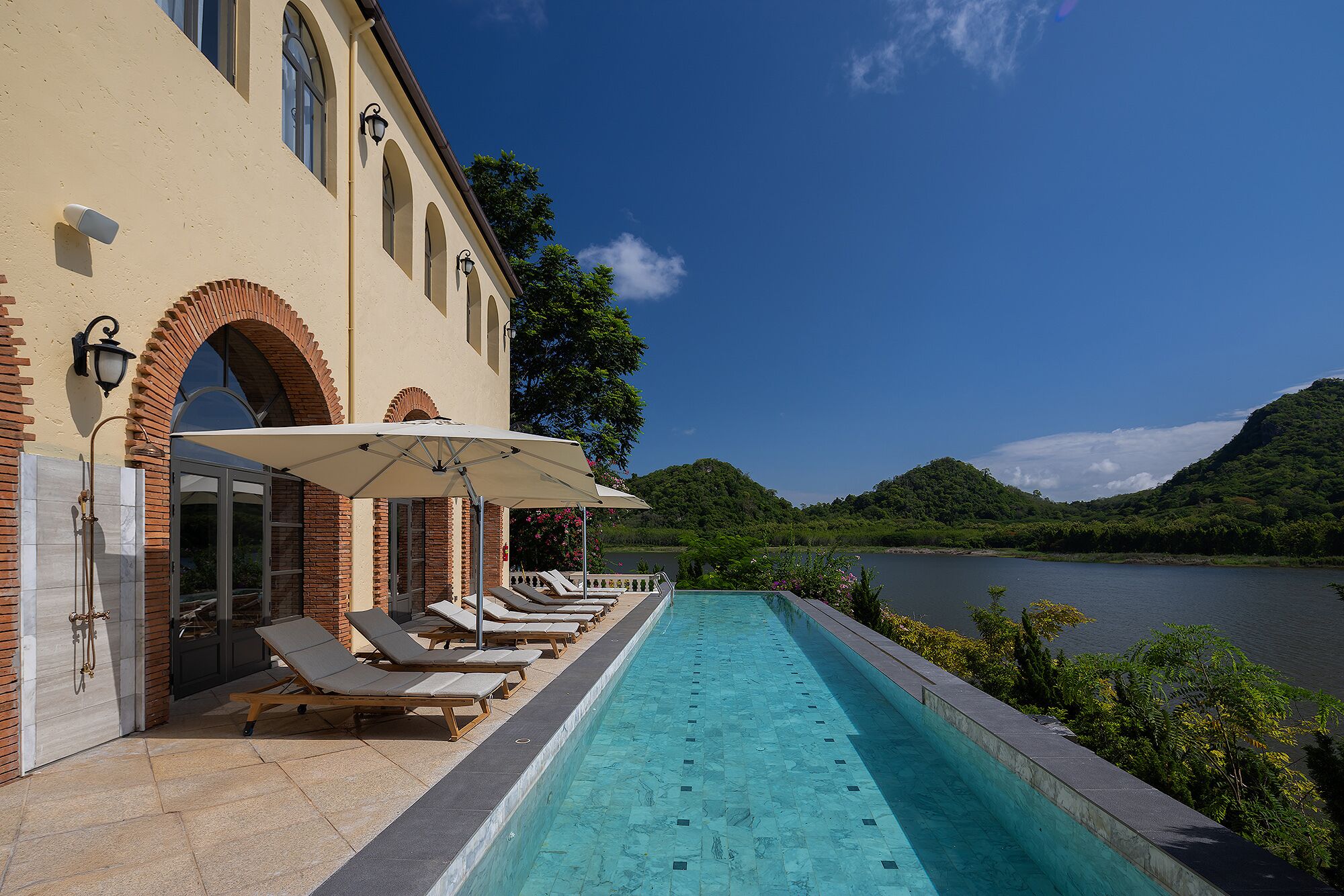 Zona de piscina con sillones de descanso y sombrillas, una piscina y vistas a un lago y colinas.