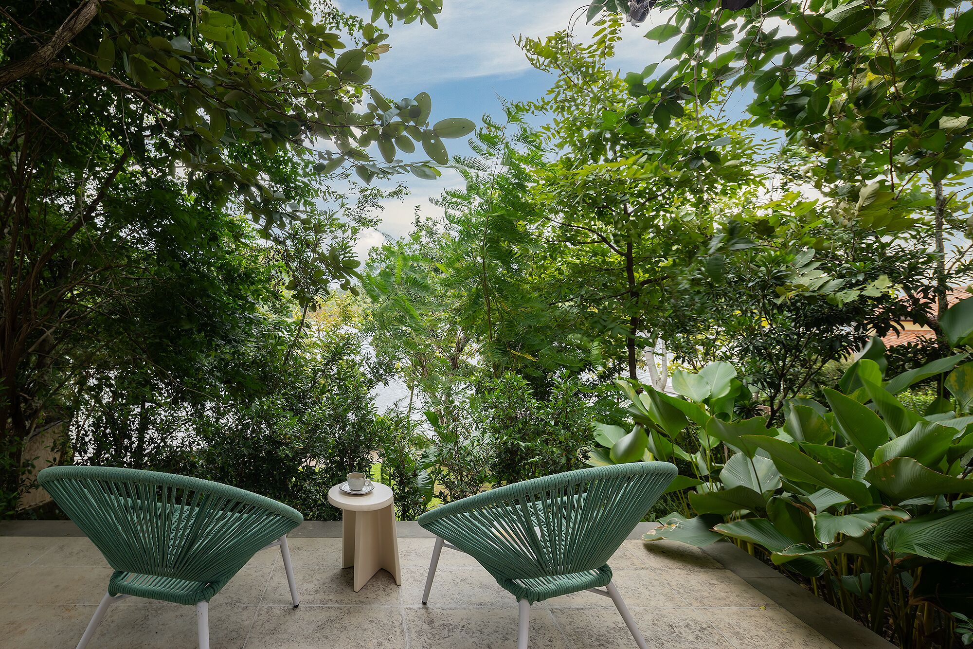 An outdoor seating area with two chairs and a table, surrounded by dense foliage and a view of the sky.