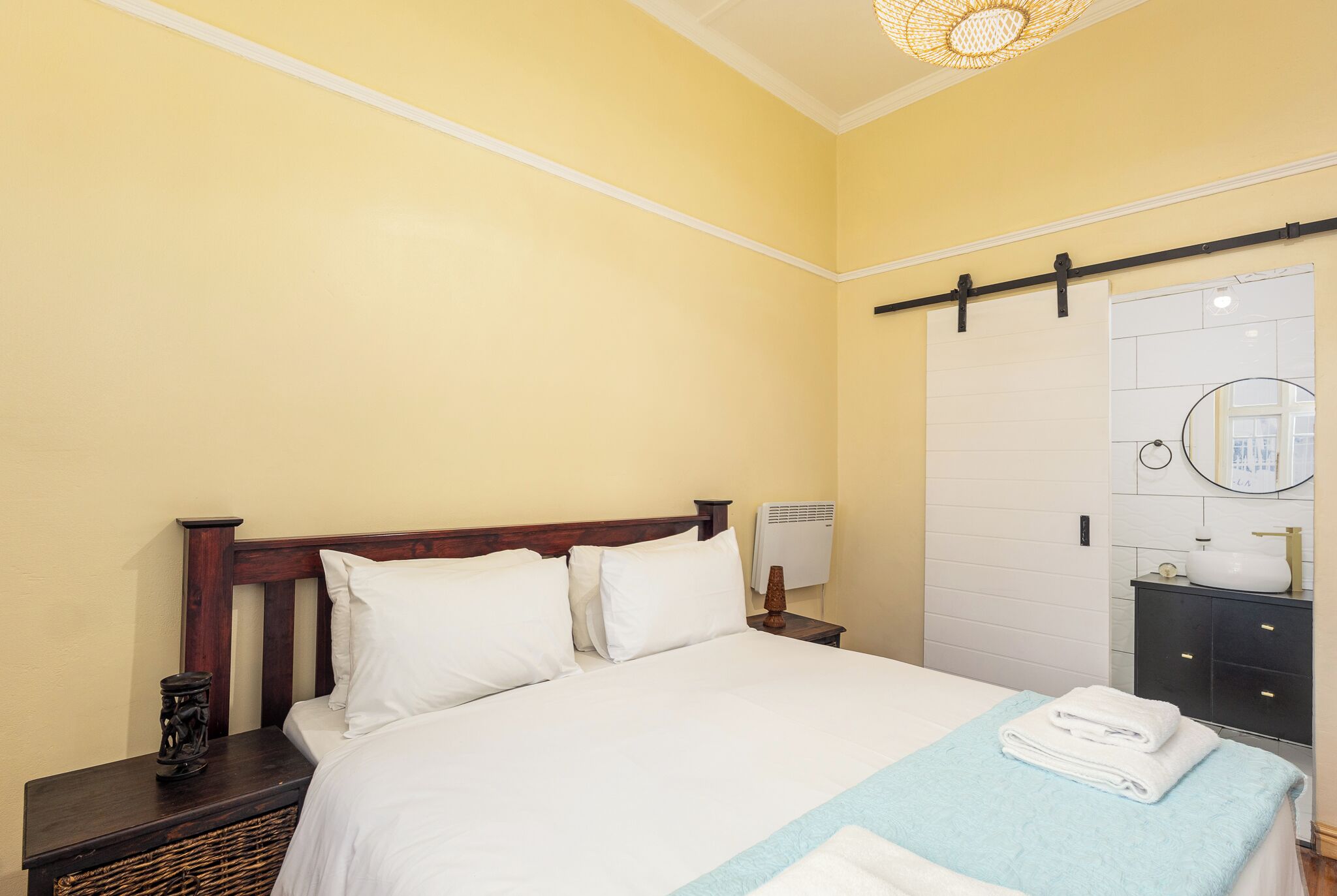 A hotel room with a bed, a nightstand, a bedside table, a wall-mounted air conditioner, a bathroom with a sink, and a towel rack.
