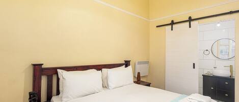 A hotel room with a bed, a nightstand, a bedside table, a wall-mounted air conditioner, a bathroom with a sink, and a towel rack.