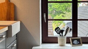 Keittiössä on ikkuna, puinen leikkuulauta, valkoinen kulho, jossa on ruokailuvälineitä, ja kehystetty kuva.