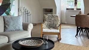 Moderni olohuone, jossa on sohva, pyöreä sohvapöytä ja nojatuoli. Huoneessa on suuri ikkuna, jossa on verhot, takka ja näkymä ulos.