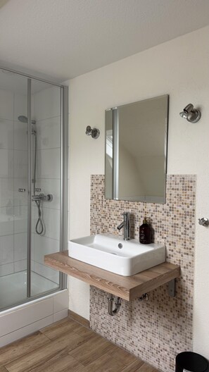Un baño con cabina de ducha de vidrio, un lavamanos blanco con encimera de madera, un espejo y salpicadero de mosaicos.