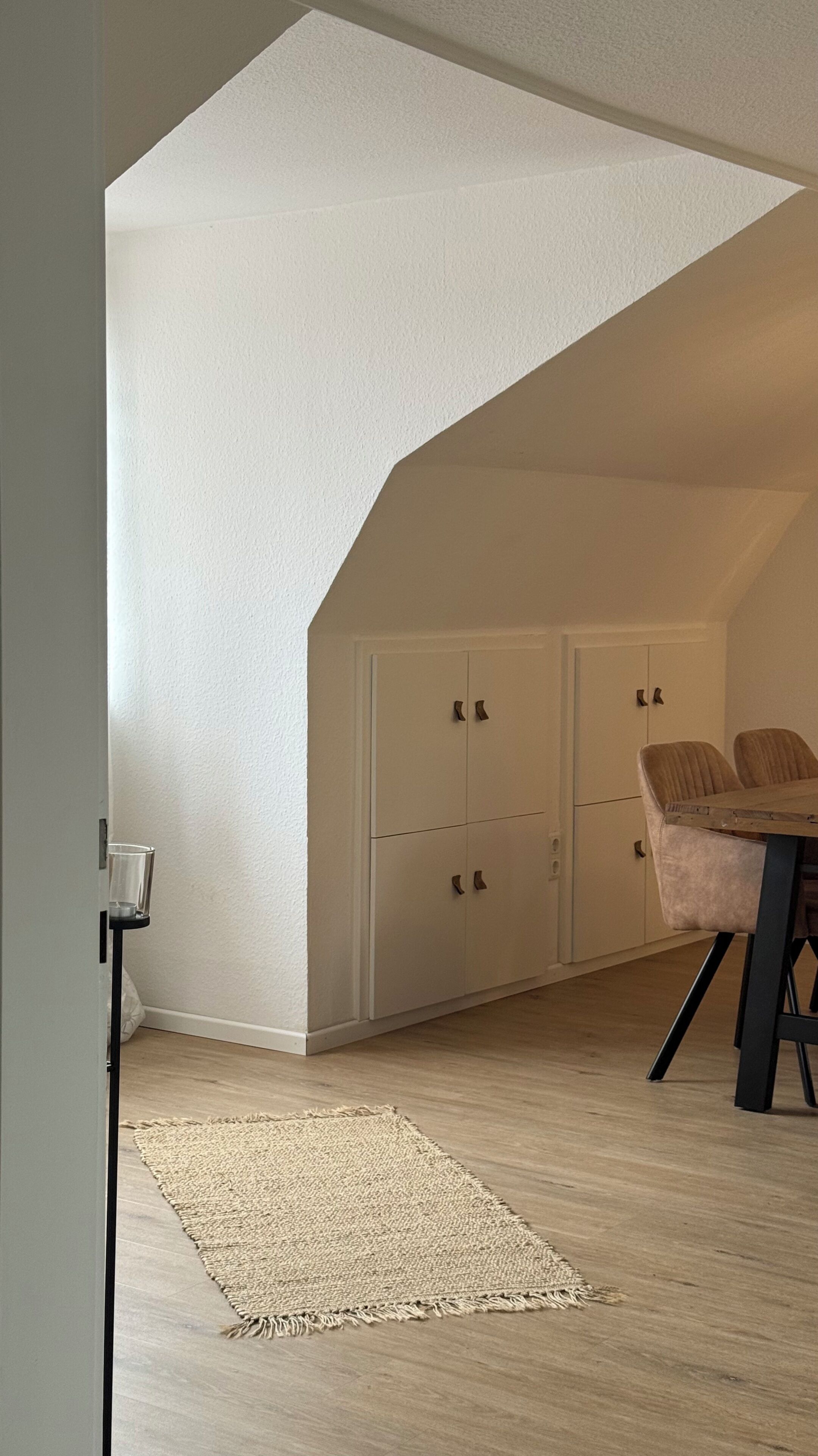 A room with a sloped ceiling, a dining table with chairs, a rug, and white cabinets with handles.
