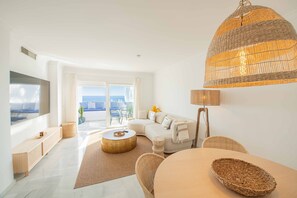 Ein modernes Wohnzimmer mit einer Couch, einem runden Couchtisch, einer Korbtischlampe und Blick durch ein großes Fenster.