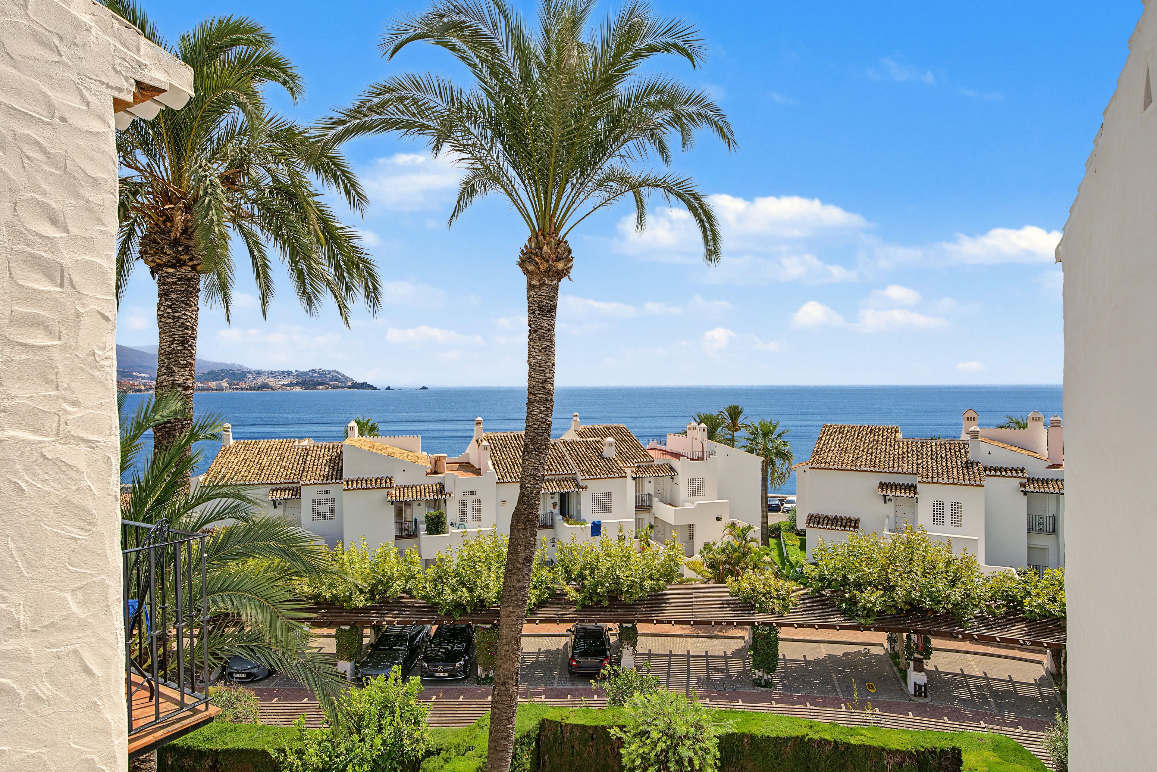 A coastal residential area with palm trees, white buildings with terracotta roofs, and a view of the sea.