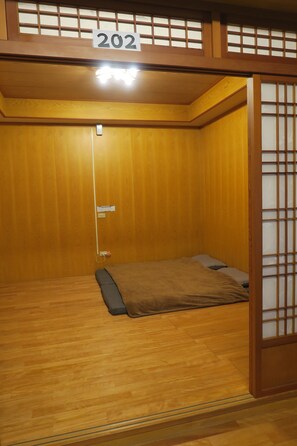 Una habitación tradicional japonesa con suelo de madera, una cama con manta marrón, una puerta con el signo de número y un celosía japonesa.