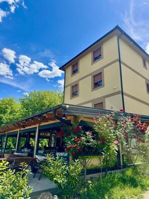 A multi-story building with a covered outdoor seating area and a garden with flowering plants. - Zvjezdana dolina Garni hotel & Glamping (Andrijevica Municipality)