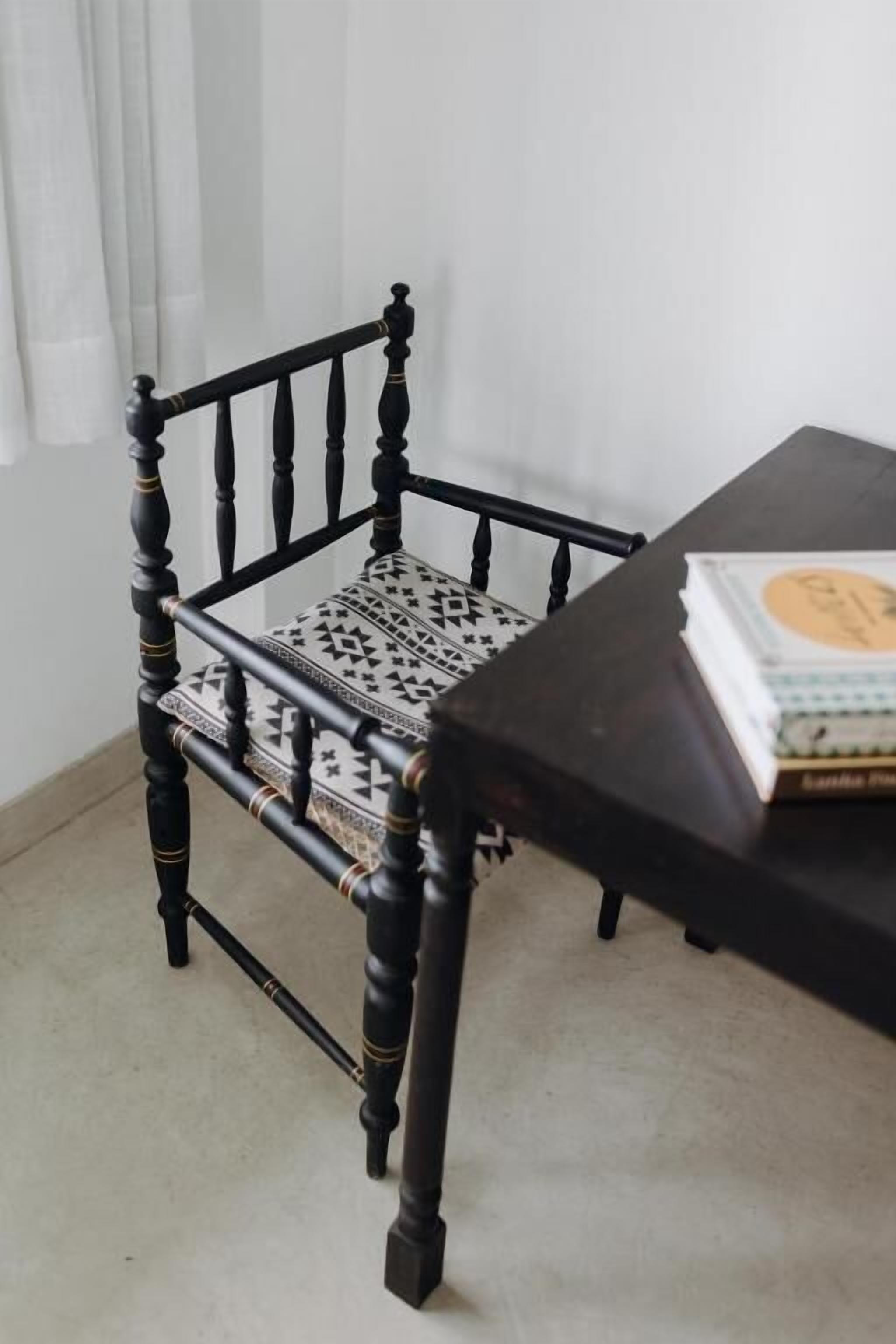 A wooden chair with a patterned cushion, a table with a book, and a white curtain in the background.
