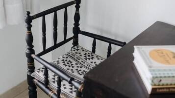 Una silla de madera con un cojín estampado, una mesa con un libro y una cortina blanca al fondo.