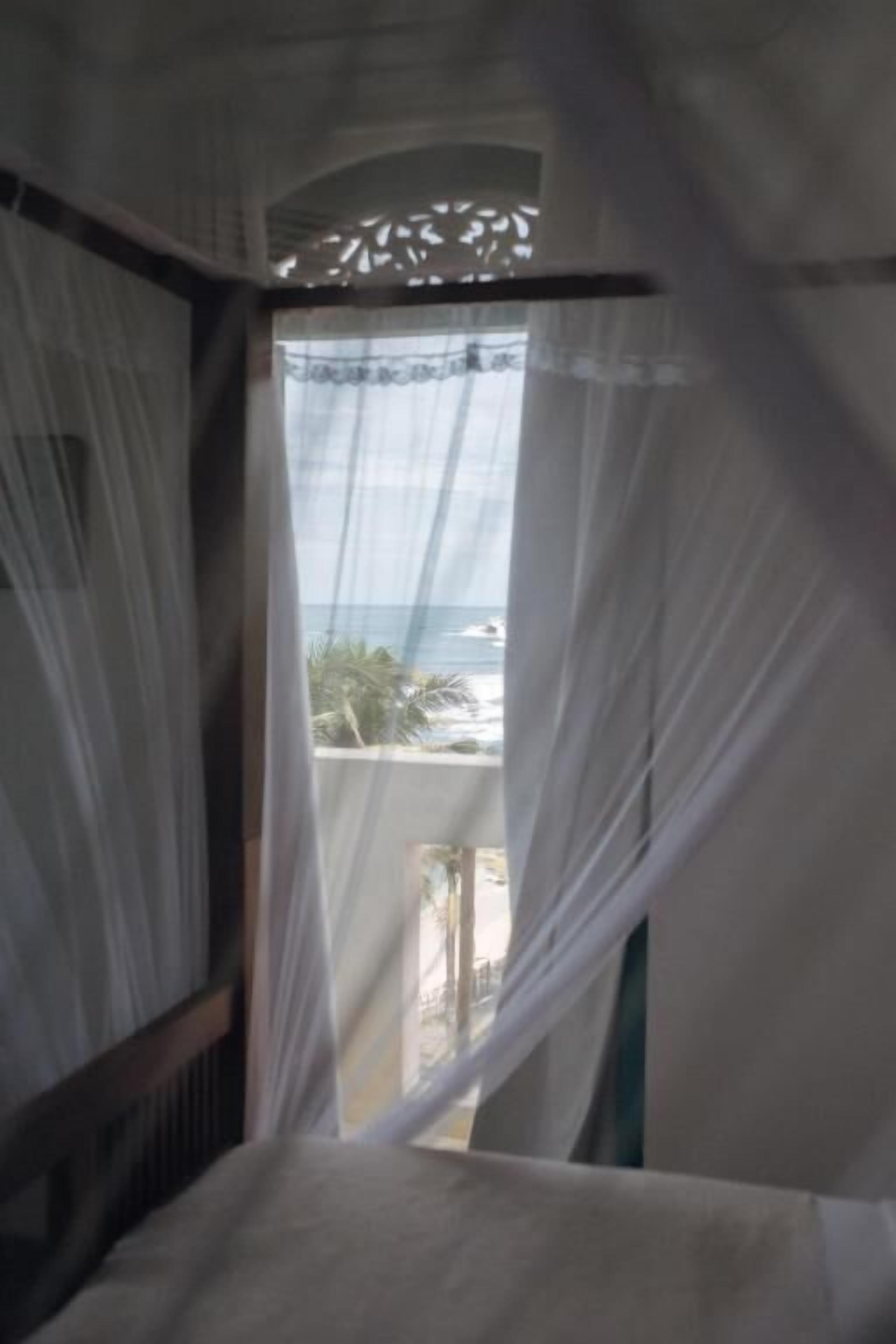 Una habitación con cama, ventana con cortinas traslúcidas y vista al mar.