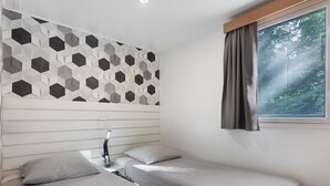 Ein Schlafzimmer mit zwei Betten, einem geometrischen Wandmuster, einem Fenster mit Vorhängen und einer Nachttischlampe.