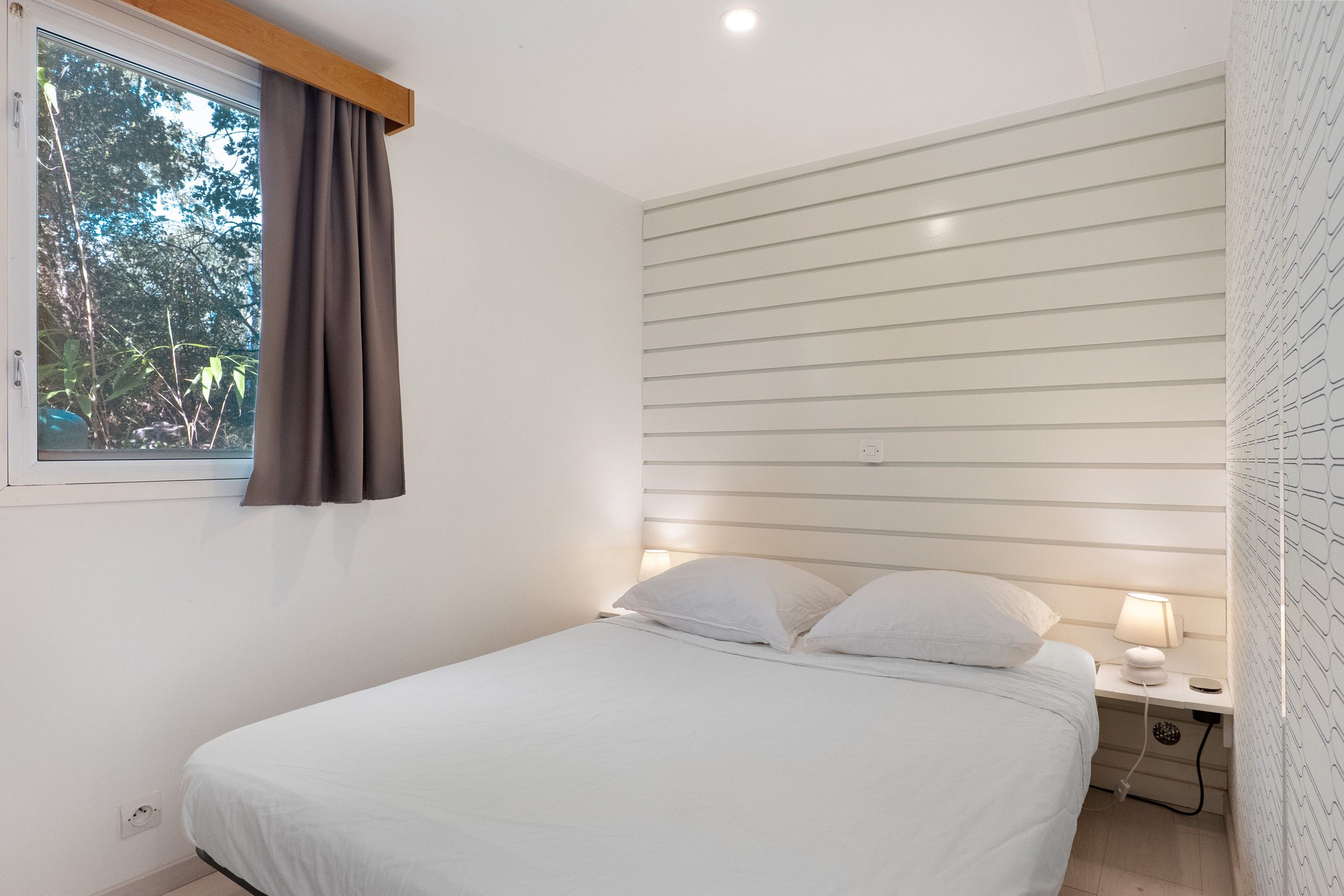 A single bed with white bedding in a room with a window, a nightstand with a lamp, and a wall with horizontal lines.