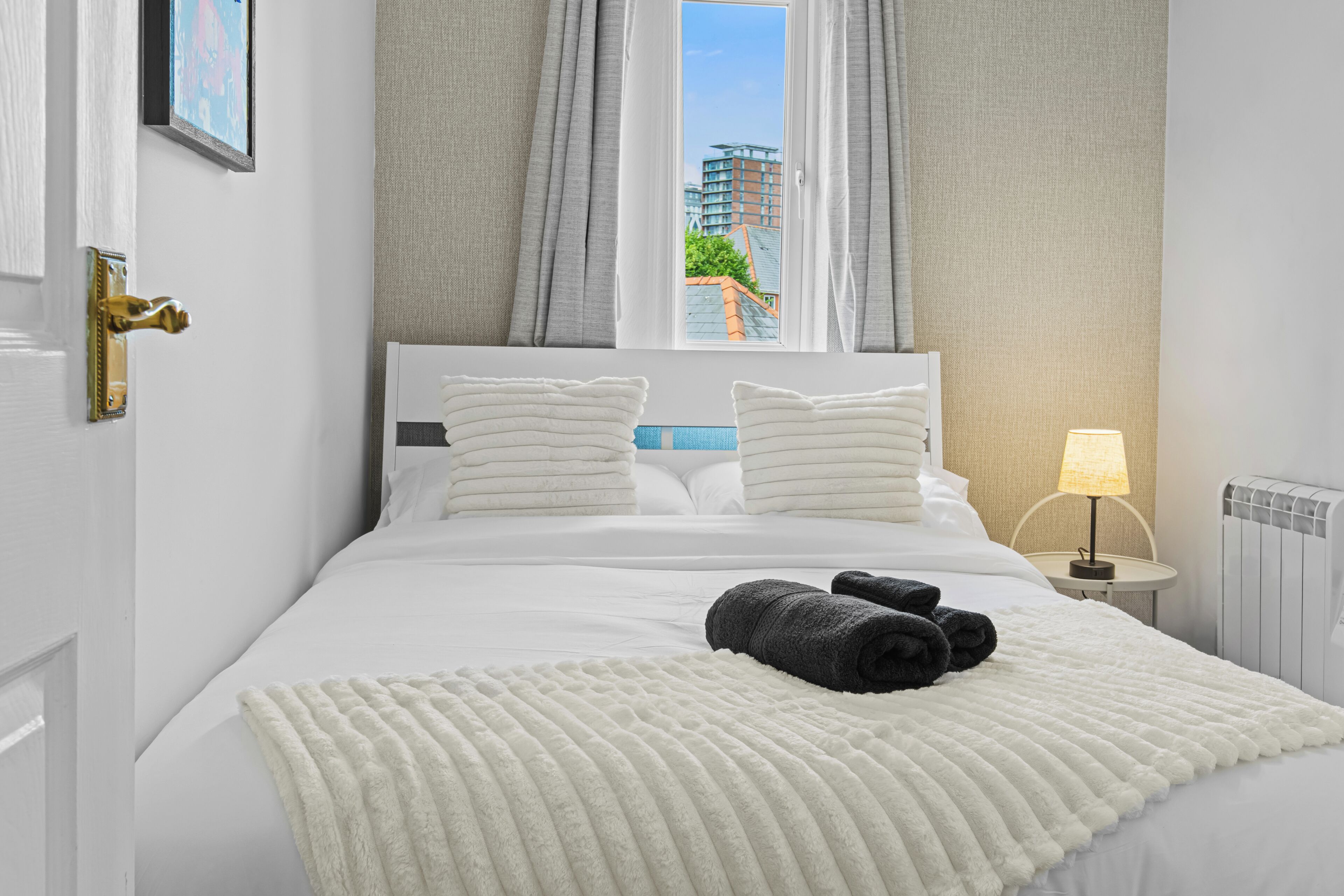 Habitación de hotel con cama, almohadas, manta, lámpara y vista a edificios a través de la ventana.