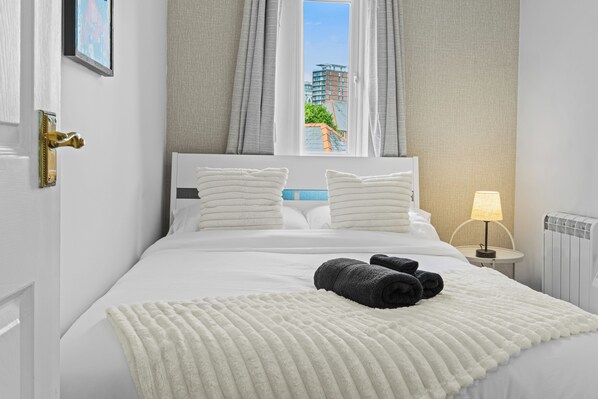 A hotel room with a bed, pillows, a blanket, a lamp, and a view of buildings through the window. - Prime location Pad/Free Parking/PS4/Wifi/Tourists (Salford)