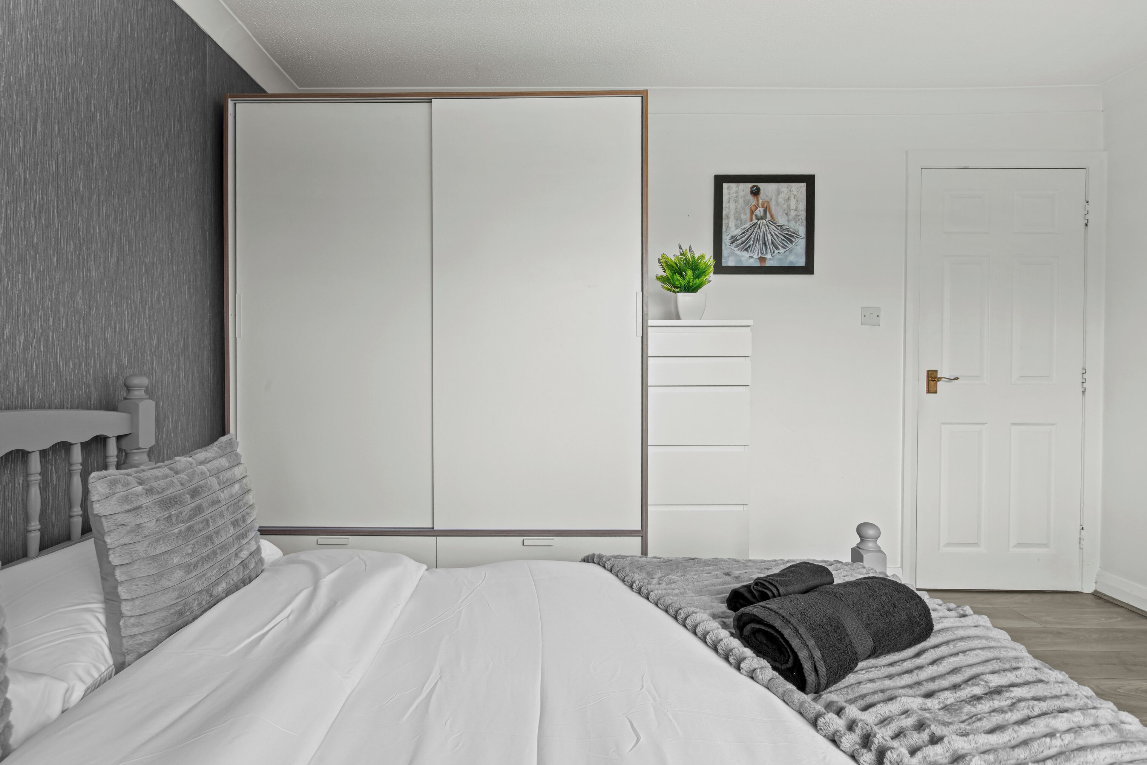 A bedroom with a bed, a wardrobe, a small potted plant, a framed picture, and a door.