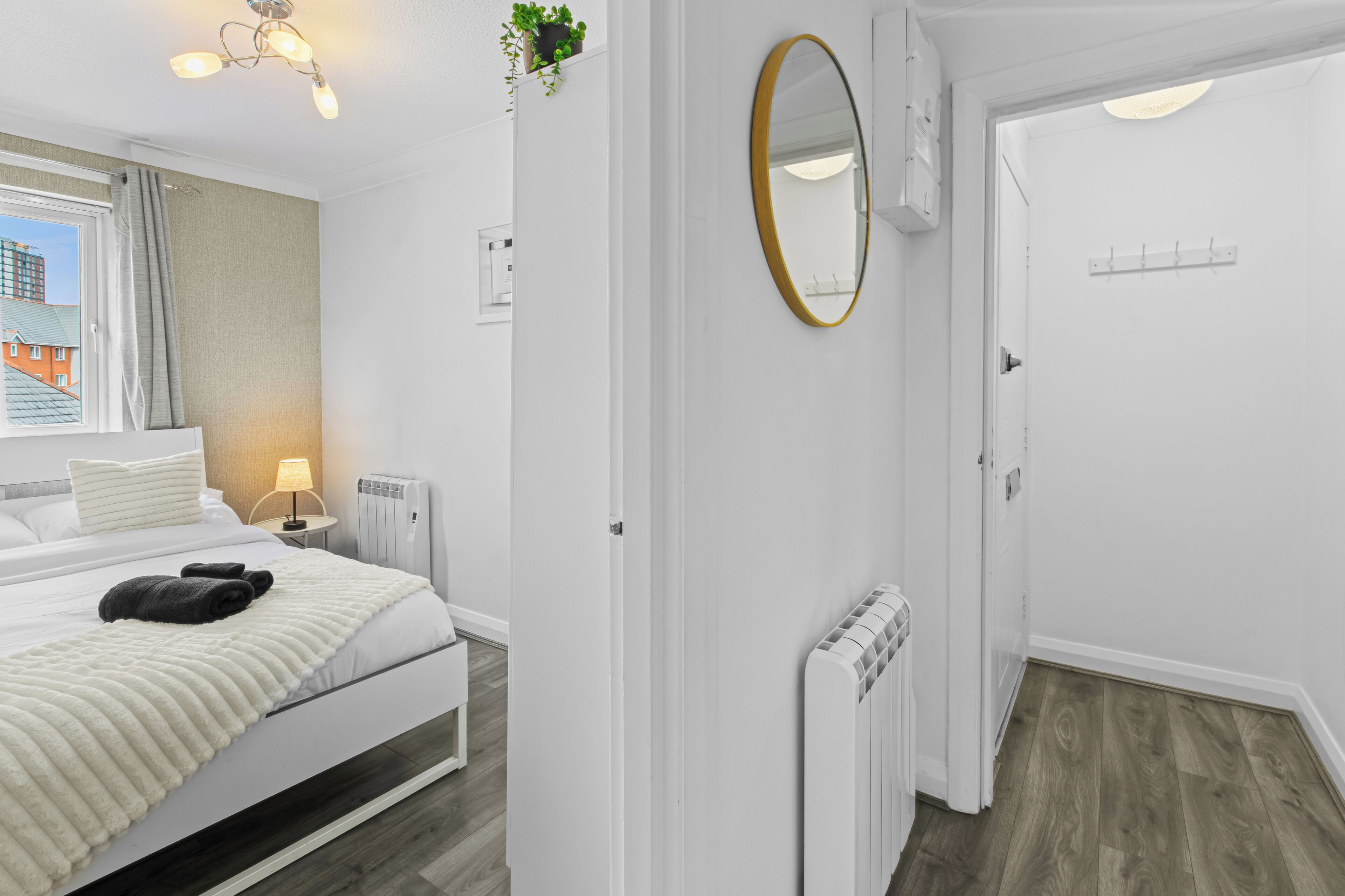 A hotel room with a bed, a bedside table with a lamp, a radiator, a mirror, and a view of buildings outside.