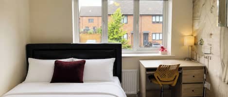 Un dormitorio con cama, escritorio, silla, radiador y ventana con vistas a edificios y árboles.