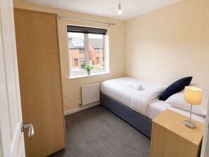A bedroom with a bed, a bedside table with a lamp, a radiator, a window with blinds, and a view of buildings outside.