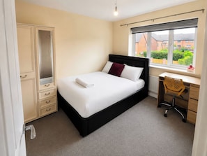 A bedroom with a bed, a desk, a chair, a wardrobe, and a window with a view of buildings.