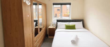 A bedroom with a bed, a wooden wardrobe, a mirror, a bedside table, and a window.