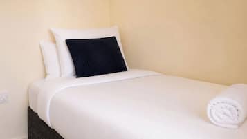 A neatly made bed with white bedding and a dark pillow, a folded white towel, and a power outlet on the wall.