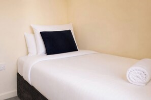 A neatly made bed with white bedding and a dark pillow, a folded white towel, and a power outlet on the wall.