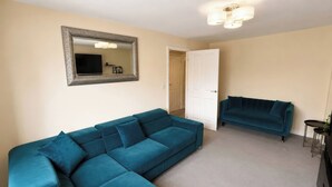 A living room with a large teal sectional sofa, a smaller teal sofa, a flat-screen TV, and a decorative mirror on the wall. - 4 Britannia Street (Salford)