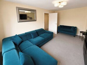A living room with a large teal sectional sofa, a smaller teal sofa, a flat-screen TV, and a decorative mirror on the wall. - 4 Britannia Street (Salford)