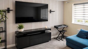 A modern living room with a large flat-screen TV mounted on the wall, a black entertainment unit, a blue sofa, and a window with blinds. - 4 Britannia Street (Salford)