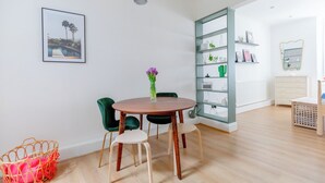 Ein Zimmer mit einem runden Holztisch, zwei grünen Stühlen, einem Korbwäscher, einem wandmontierten Regal mit Dekoration und einem gerahmten Bild.