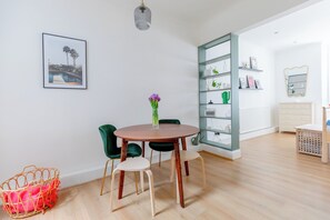 Ein Zimmer mit einem runden Holztisch, zwei grünen Stühlen, einem Korbwäscher, einem wandmontierten Regal mit Dekoration und einem gerahmten Bild.