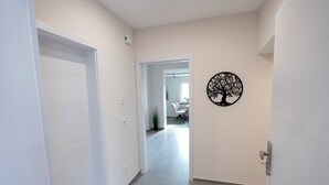 Un couloir avec une porte blanche, une décoration en forme d’arbre fixée au mur, et une vue sur une pièce où l’on aperçoit une chaise et une plante.