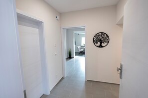 Ein Flur mit einer weißen Tür, einer Wanddekoration mit Baummotiv und Blick in einen Raum mit Sessel und Pflanze.