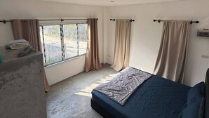 Una habitación con cama, ventana con cortinas, un bloque de concreto y una lámpara en el techo.