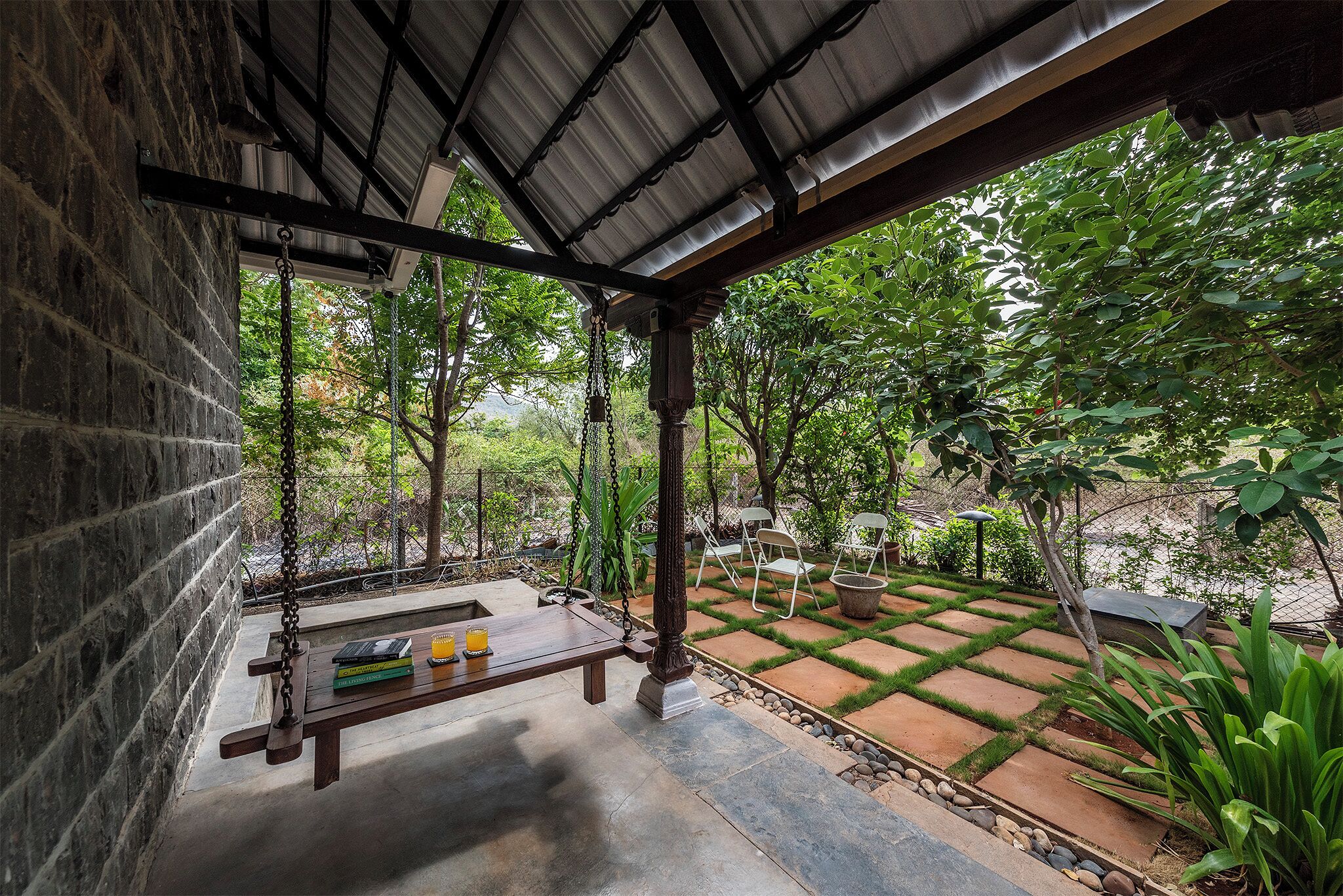 A covered patio with a swing, a table with books and glasses, and a garden with a stone pathway and chairs.