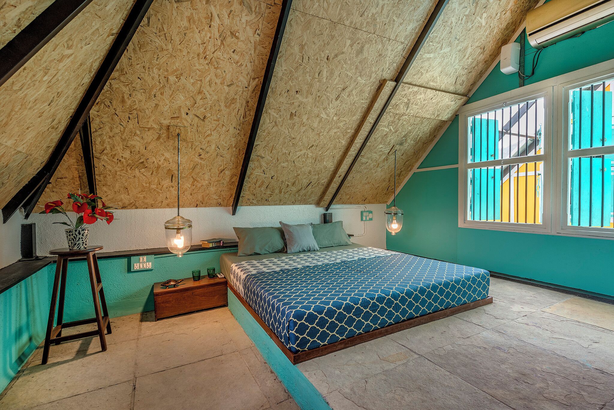 Ein Schlafzimmer mit schrägem Dach, einem Bett mit gemustertem Überwurf, einem Nachttisch mit Vase und einer Wandleuchte.