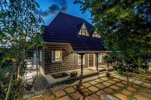 A stone house with a dark roof, a porch, and a small garden area. - CosmicStays Raanjai  (Mulshi)
