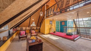 Um amplo loft com vigas de madeira aparentes, mezanino com grade de ferro forjado, cadeira balanço e um banco vermelho.