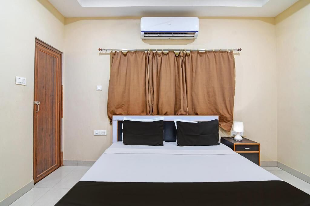 A bedroom with a bed, a nightstand, a lamp, an air conditioner, a door, and curtains.