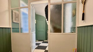 Una escala amb un cubell metàl·lic a la dreta, una porta que dona a un espai amb terra de pissos i un rellotge de paret sobre la porta.